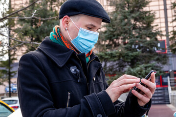 The young man with a face mask on, trying to make a video call on his smartphone. Outdoors. Coronavirus protection measures.