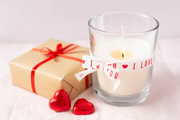 The theme is Valentine's Day. A burning candle and two red candy in the shape of hearts and packed gift are on a white background. Free space for text.
