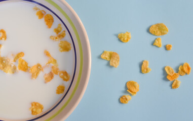 Pieces of cornflakes spread around on a light blue surface next to a bowl of milk with cornflakes