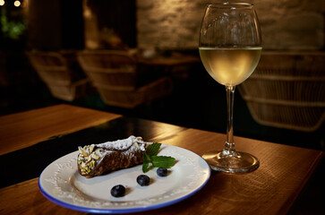 Sicilian cannoli roll with blueberries on a white ceramic plate and a glass of misted white wine on a restaurant table with a blurred background.
