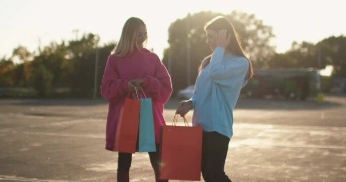 Happy girl friends look at purchases and hug smiling. Bags with clothes and accessories after shopping. Empty parking lot with trees at sunset