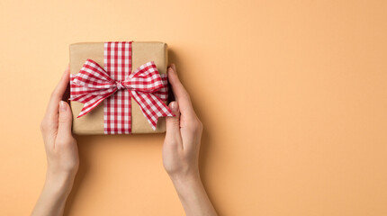 First person top view photo of saint valentine's day decorations female hands giving craft paper giftbox with checkered ribbon bow on isolated beige background with empty space