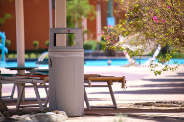 Empty deck chairs under straw shade umbrellas on swimming pool side and litter bin for garbage...