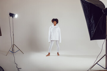 African American woman with puffy hair in a white hoodie and leggings stands on a white background in a photo studio.