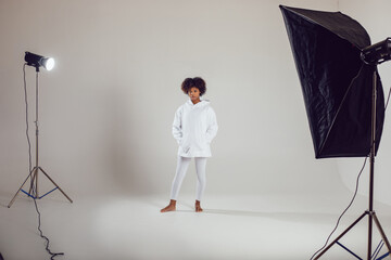 African American woman with puffy hair in a white hoodie and leggings stands on a white background...