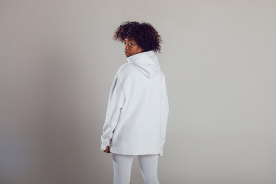 African American woman with puffy hair in a white hoodie and leggings stands on a white background in a photo studio.
