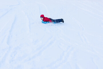 Boy sliding on blue tubing on stomach in winter clothes among white snow, distant view, copyspace. Inflatable sleigh rides.