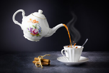 Levitating teapot pouring tea into cup with steam, cinnamon tied with rope, on gray table on dark...