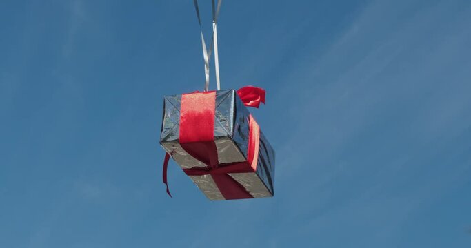 Silver helium balloons attached to present box float in air against blue sky. Glossy case tied with red ribbon reflects bright sunlight closeup