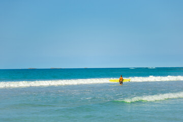 Beautiful seascape with a surfer with a yellow surfboard. Sports recreation at sea. Coastal blue sea waves under a blue sky on a sunny day.