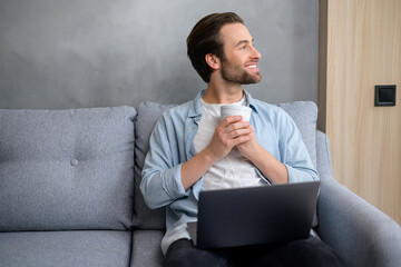 Charismatic caucasian man sitting down on the sofa with a laptop on his laps, looking away and smiling. Freelancer man enjoys remote work while drinking coffee