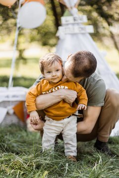 Caring young father hugging and kissing his little son while posing together outdoors. Blur background of colorful balloons and toy teepee tent.