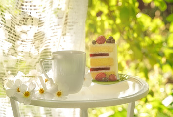 Beautiful porcelain coffee cup and slice of cake on glass table decorated with white flowers in summer garden in sunlight. Copy space. Summer drinks concept.