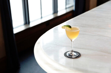 A yellow colored cocktail with a lime slice on the edge of a white granite table surface in a restaurant. 
