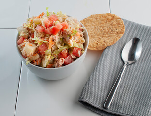 Tuna salad in a white bowl with lettuce pasta mayo carrots and red peppers on top with a 
 tostada and a spoon on the side. Horizontal