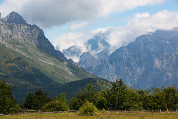 View of the rocky peaks of the Pyrenees, covered with low clouds. In the foreground is a fenced green meadow and trees.