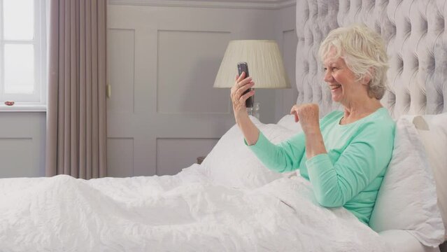 Senior woman at home wearing pyjamas in bed enjoying video call with family on mobile phone - shot in slow motion