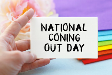 A woman's hand holds a piece of paper with the inscription NATIONAL Coning Out DAY, against the background of a stack of cloth folded in the colors of the LGBT flag. LGBT concept.