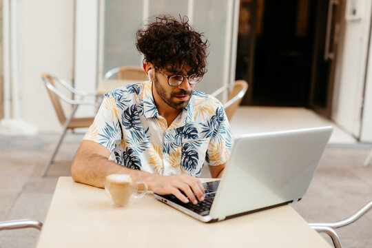 Young man working on his business at a table with chairs outside a coffee shop. He's typing on his laptop and has a latte on the table.