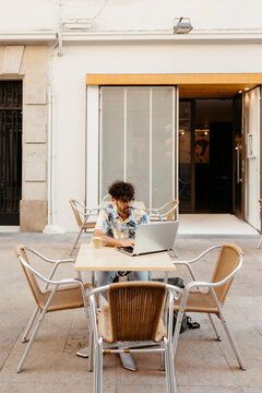 Young man working on his business at a table with chairs outside a coffee shop. He's typing on his laptop and has a latte on the table.
