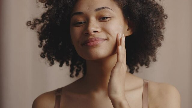 Female beauty procedures. Young attractive african american lady applying smear of pampering cream on her cheek