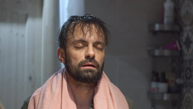 A man in the bathroom in front of a mirror sleepily looks at his reflection with a towel around his neck
