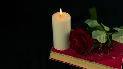 A red rose with a book and a burning candle on a black background. From above of burning candle with fresh red with a rose on book on black background