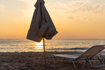 Evening on the beach. A closed beach umbrella obscures the setting sun. A chaise lounge on the...