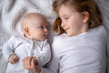 Happy children, toddler and older sister, hugging at home on a white blanket, smiling, shot from above. blonde girl hugging her newborn sister