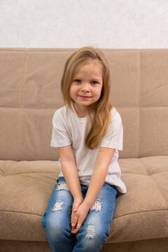 Portrait of a sad little blonde girl in a white T-shirt. Sitting on the couch unhappy
