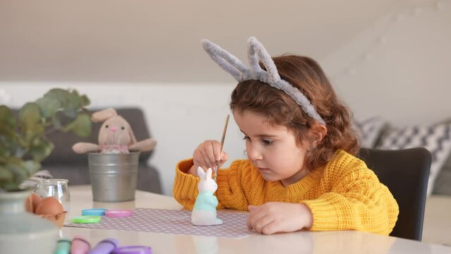 Curly girl prepares Easter decoration by painting a clay bunny and eggs. Easter celebration spring vacation and party event