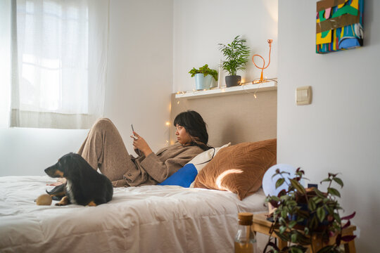 Asian Woman Laying On The Bed And Reading With Her Dog.