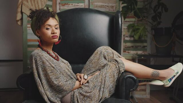 Young confident stylish african american lady looking at camera sitting in black leather armchair, tracking shot