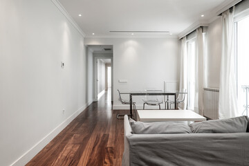 Living room with long gray sofa, glass dining table and perspex chairs with reddish wood flooring and soft gray painted walls