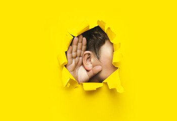 Close-up of a man's ear and hand through a torn hole in the paper. Yellow background, copy space. The concept of eavesdropping, espionage, gossip and tabloids.