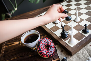 Fototapeta na wymiar hand holds a white vintage chess piece. Chess board and scattered chess on wooden table. Chocolate doughnut for morning breakfast. board game Concept