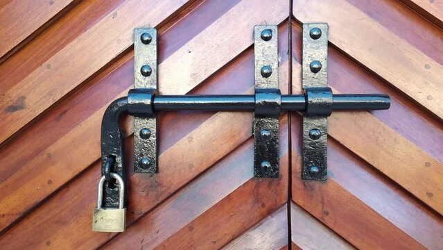 Metal security bar on a wooden double door with padlock. Black steel latch on brown heavy carved gate with fishbone pattern - close up.