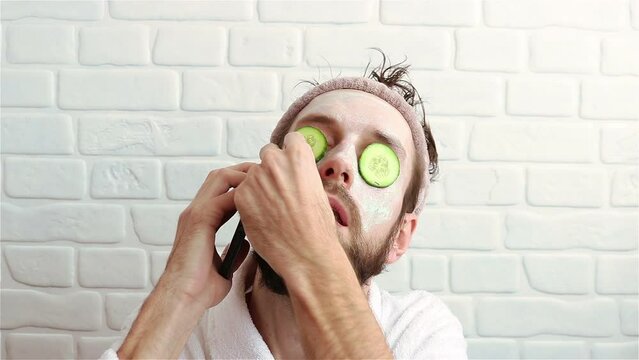 bearded man with facial cosmetic mask and cucumber slices calling phone funny video