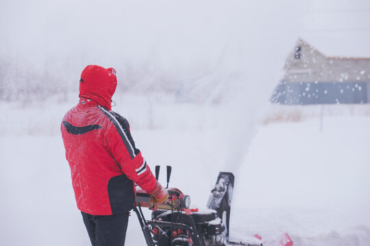 Man (view From The Back) In Windproof Cleans, Clears Snowdrifts And Drifts In Blizzard Outside The City. Particles Of Snow And Drifts Fly Everywhere. Selective Focus, Inclement Weather, Copy Space.