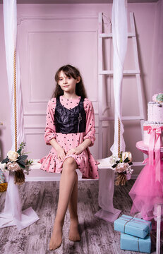 Portrait of a cute little girl sitting on a hanging swing with gifts in her hands. Beautiful baby in a dress on a pink studio background.