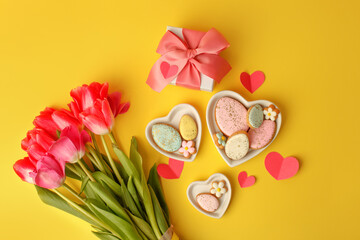 Sweets, pastry, gingerbread cookies for Easter table. Easter eggs heart shaped decor plate, pink tulips on yellow background top view copy space, spring seasonal holiday banner for site, flyer, coupon
