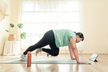 Asian young female plus size in sport wear effort doing exercise on yoga mat in living room. Watching training in loss weight course online video in tablet. Healthy lifestyle concept.