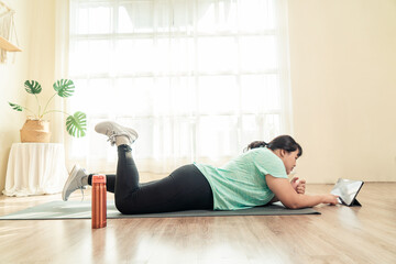Asian young female plus size in sport wear lying tired from exercise training on yoga mat in living room. Training in loss weight course online video in tablet. Healthy lifestyle concept.