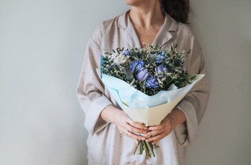 Woman in home clothes with bouqet of blue and lilac flowers in hands on grey background