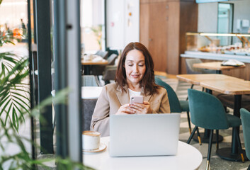 Adult smiling brunette business woman forty years with long hair in stylish beige suit working on laptop drinking coffee at the cafe