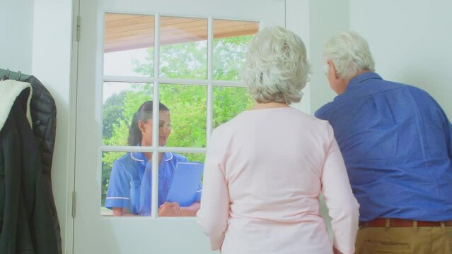 Senior couple at home greeting female care worker in uniform making home visit at door - shot in slow motion