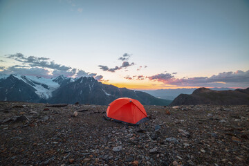 Scenic alpine landscape with tent at very high altitude with view to large mountains in orange dawn sky. Vivid orange tent with awesome view to high mountain range under cloudy sky in sunset colors.