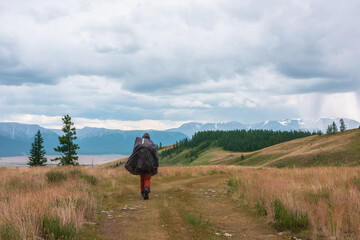 Fototapeta na wymiar Tourist walks through hills and forest towards bad weather. Hiker on way to large snow mountain range under rainy cloudy sky. Man in raincoat in mountains in overcast. Traveler goes towards adventure.