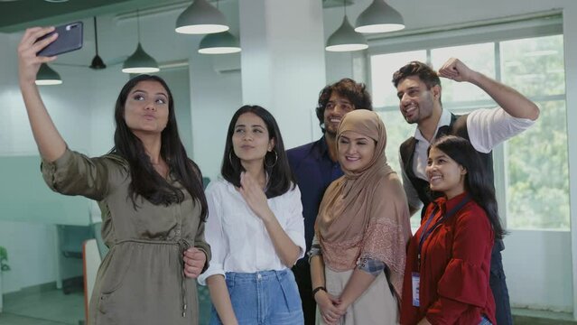 Happy Indian group of young office employees taking a selfie on cellphone in a modern office space. Asian team of cheerful and mixed gender associates taking a selfie picture or photo on a occasion.