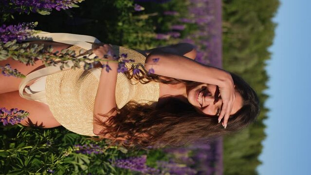 An attractive brunette woman stands in a white blouse in the middle of a field of lupines. Covered by a straw hat and smiling. Sweet emotions. Vertical video